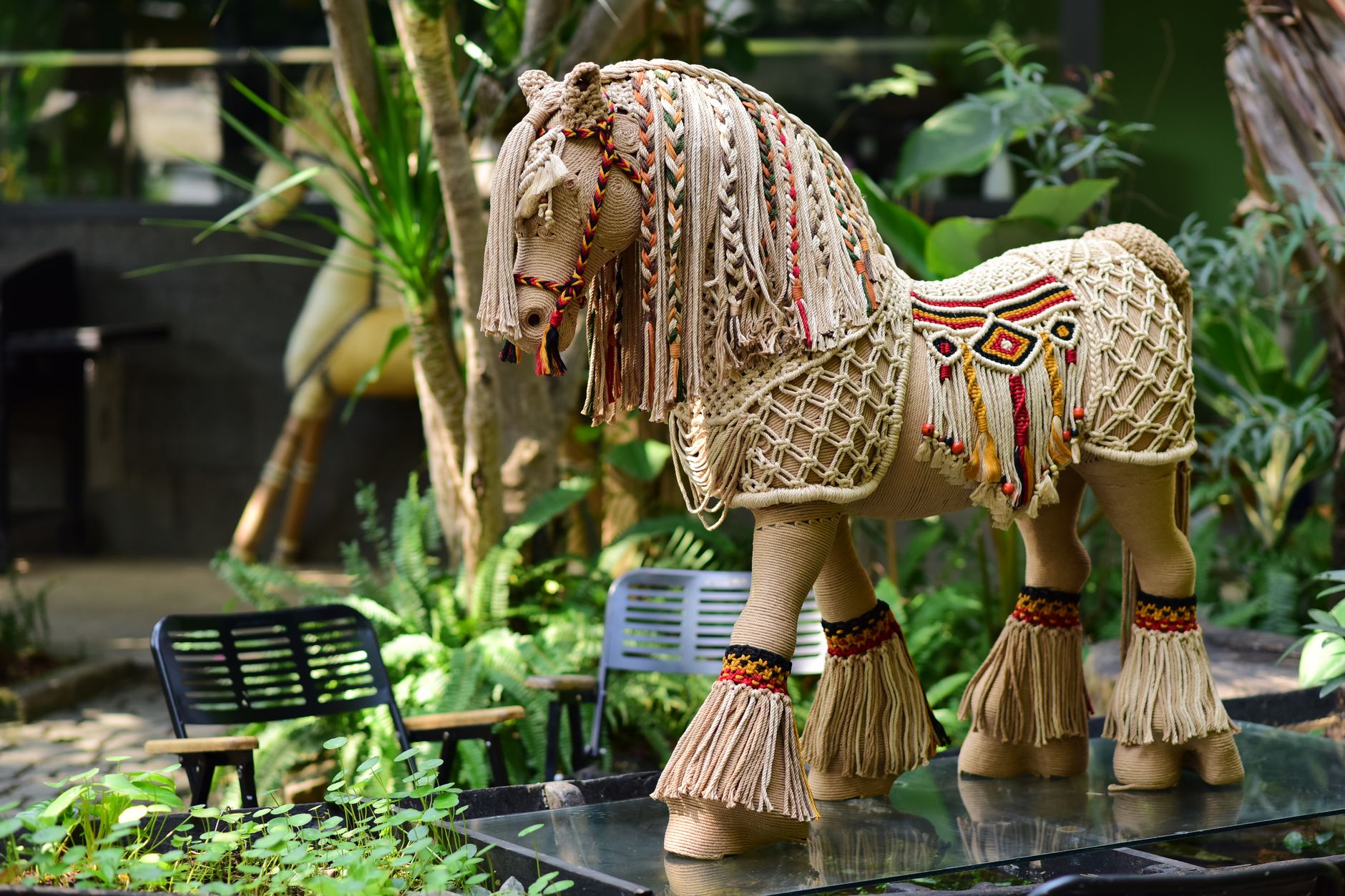 Intricately woven horse sculpture outdoors surrounded by lush greenery and vibrant chairs.