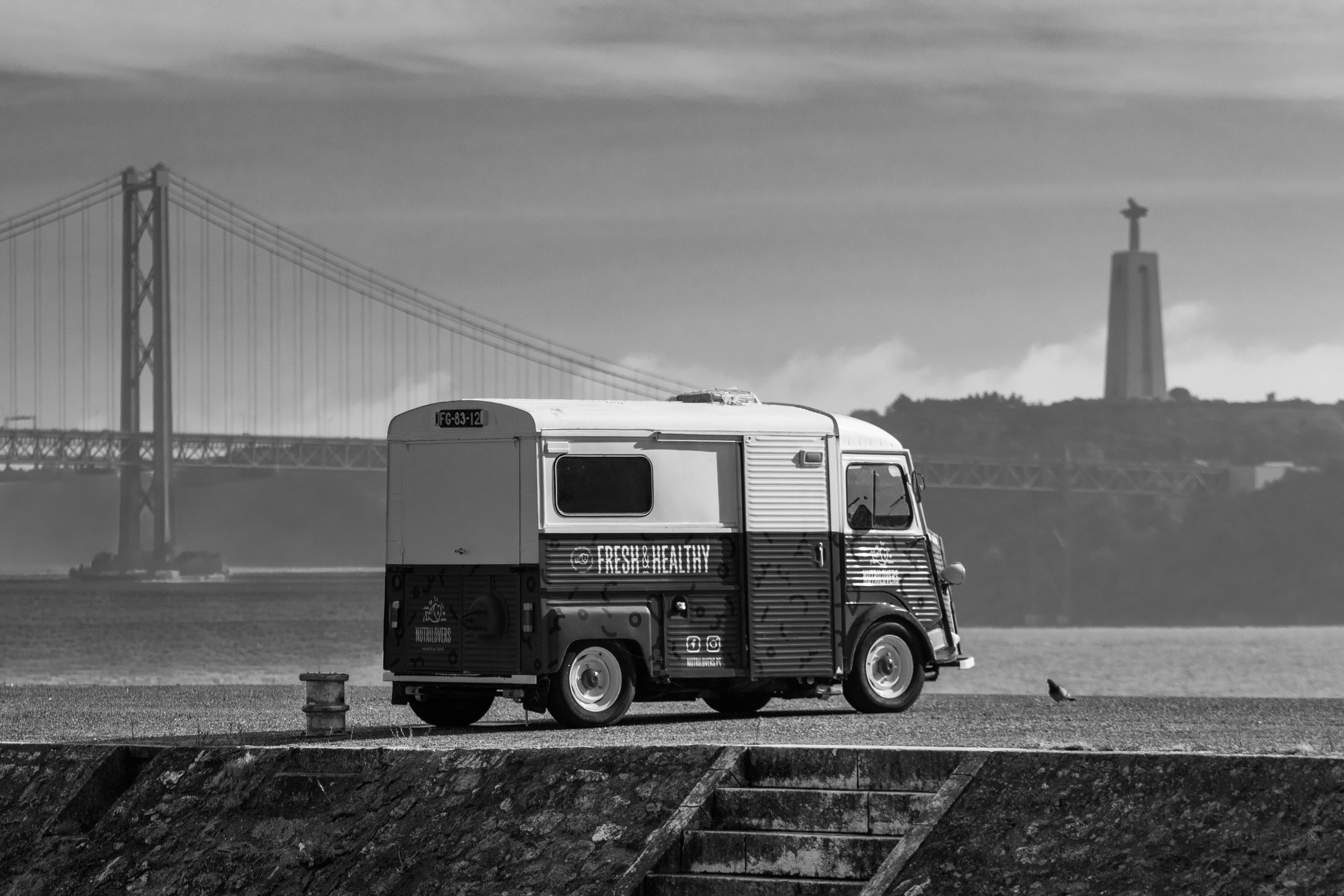 Black and white photo of a vintage van near the Lisbon Bridge and Christ Statue, capturing the essence of Lisbon.