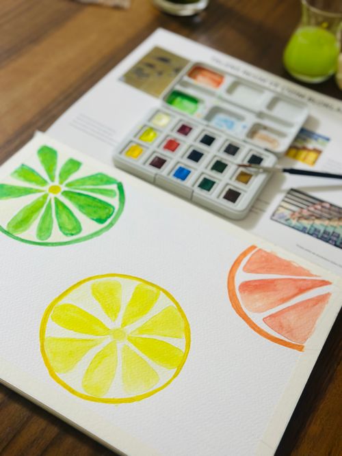 Free Artistic watercolor citrus slices in green, yellow, and orange tones on paper with paint palette. Stock Photo