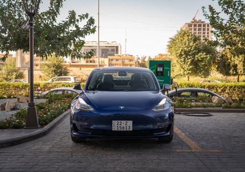 Free A blue Tesla Model 3 charges at an outdoor station during the day, surrounded by greenery and urban architecture. Stock Photo