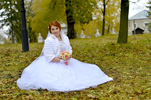 Bride in a white wedding dress sitting outdoors in an autumn park holding a bouquet.