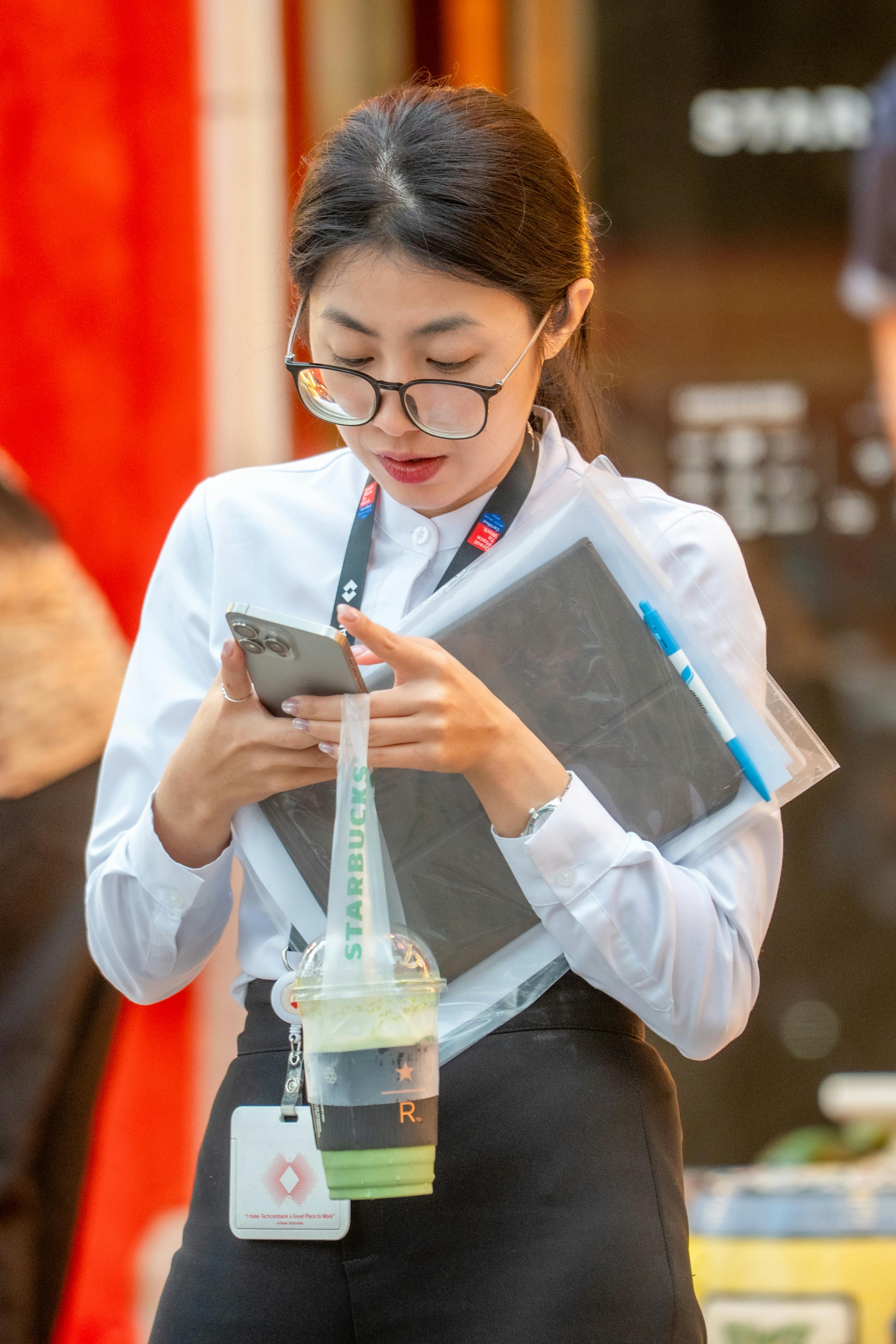бесплатная Деловая женщина проверяет телефон, держа в руках напиток и планшет, на открытом воздухе. Стоковое фото
