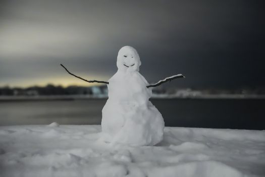 A playful snowman stands by a serene lakeside, silhouetted against the winter dusk.