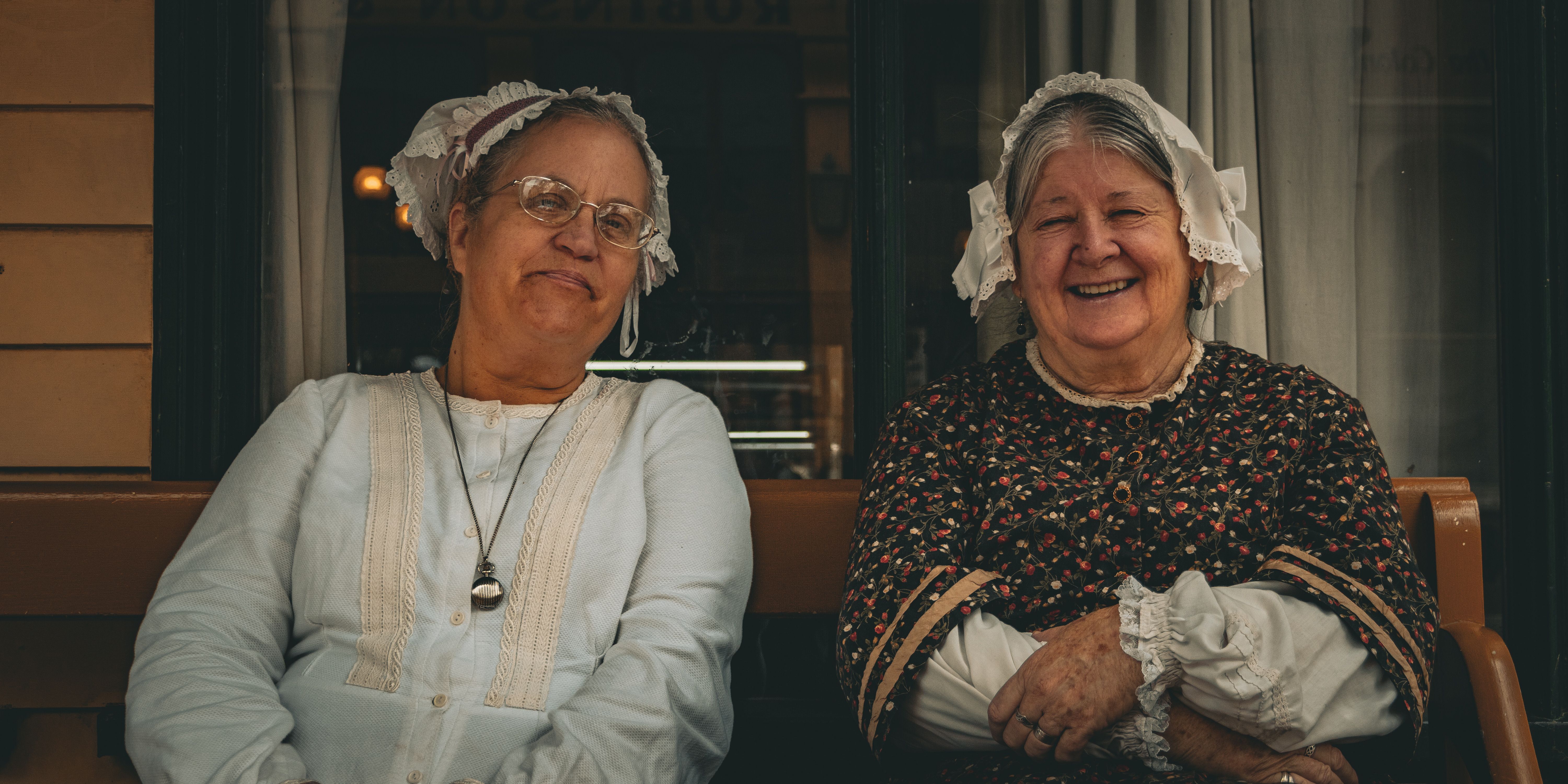 Two senior women in vintage attire smiling, seated outside a building.