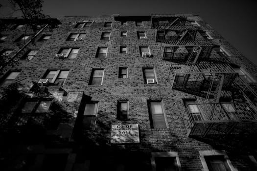 Black and white image of a brick building with fire escapes, for sale sign visible.