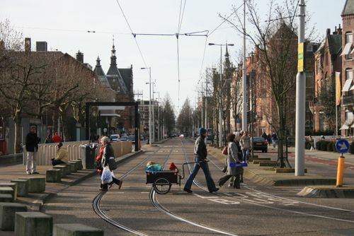 Free Street scene with people crossing tram lines in a European city, flanked by historic buildings. Stock Photo