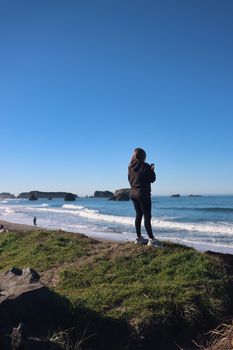 A woman takes a photo of the ocean from a grassy cliff under a clear blue sky.