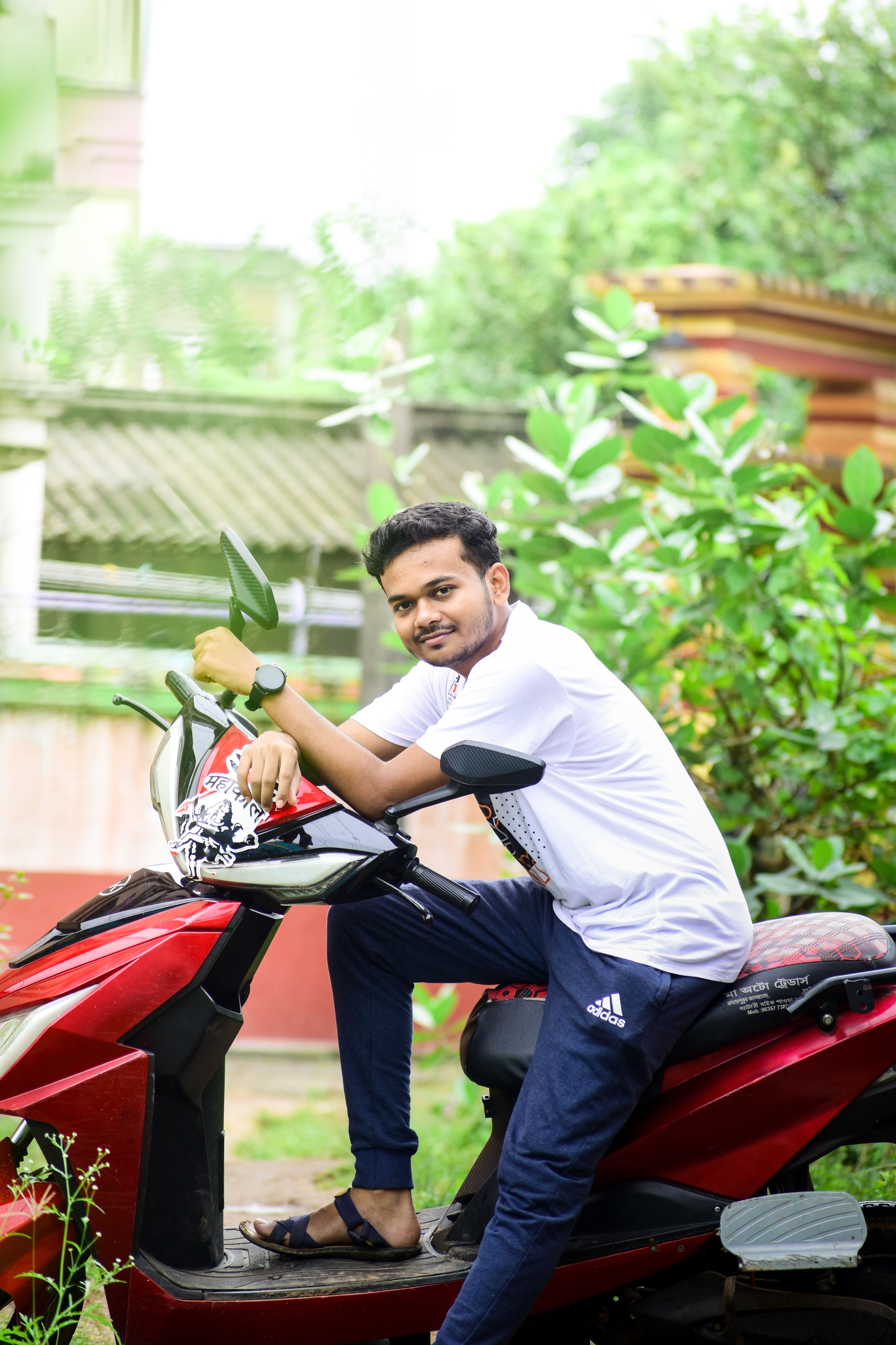 Smiling young adult male sitting on a red scooter with a lush garden background.