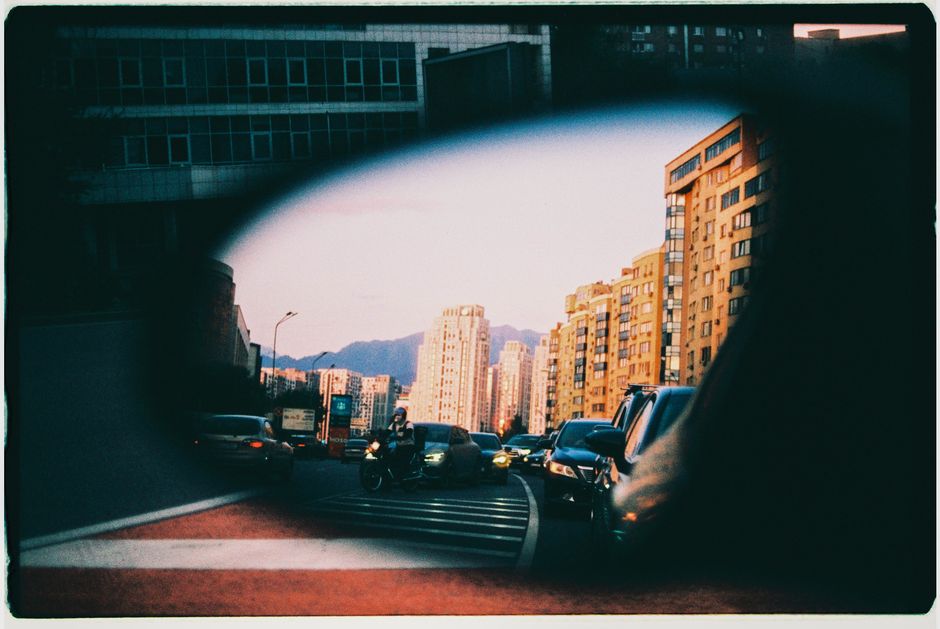 Urban reflection in car mirror with city buildings and mountains at sunset in Almaty.