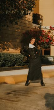 Stylish woman poses in a black outfit with vibrant flowers at sunset in an urban environment.