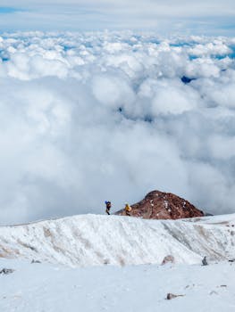 Two hikers exploring a snowy mountain peak surrounded by clouds and sky.