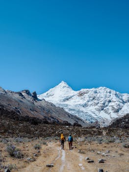 Two hikers traverse a rugged path in the Andes mountains under a vibrant blue sky.