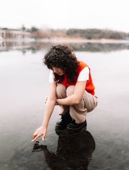 A young woman crouches by a serene lake, gently touching the water's surface during a tranquil day.
