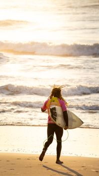 A surfer in a wetsuit runs towards the waves, carrying a surfboard along a sunny beach.