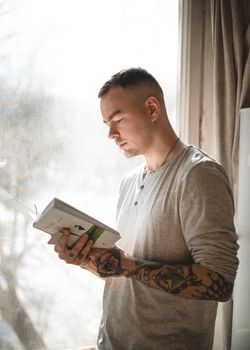A young tattooed man enjoys a book near a sunlit window, creating a serene reading moment.