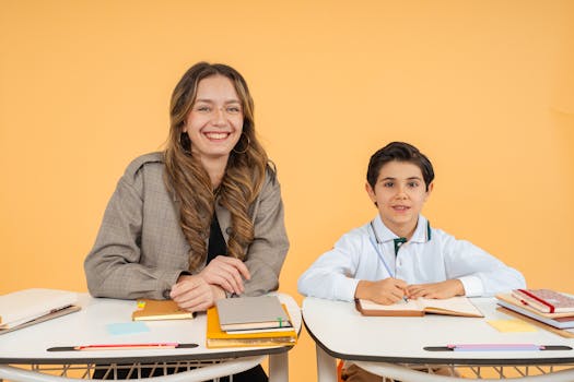 A teacher and student sitting at desks with books and notebooks, smiling warmly.