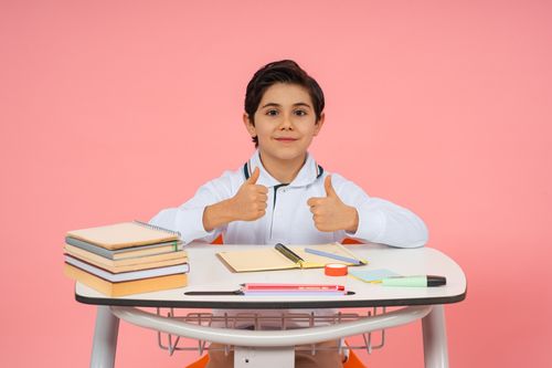 Free A happy schoolboy giving thumbs up while seated at a desk with school supplies, conveying positivity in education. Stock Photo