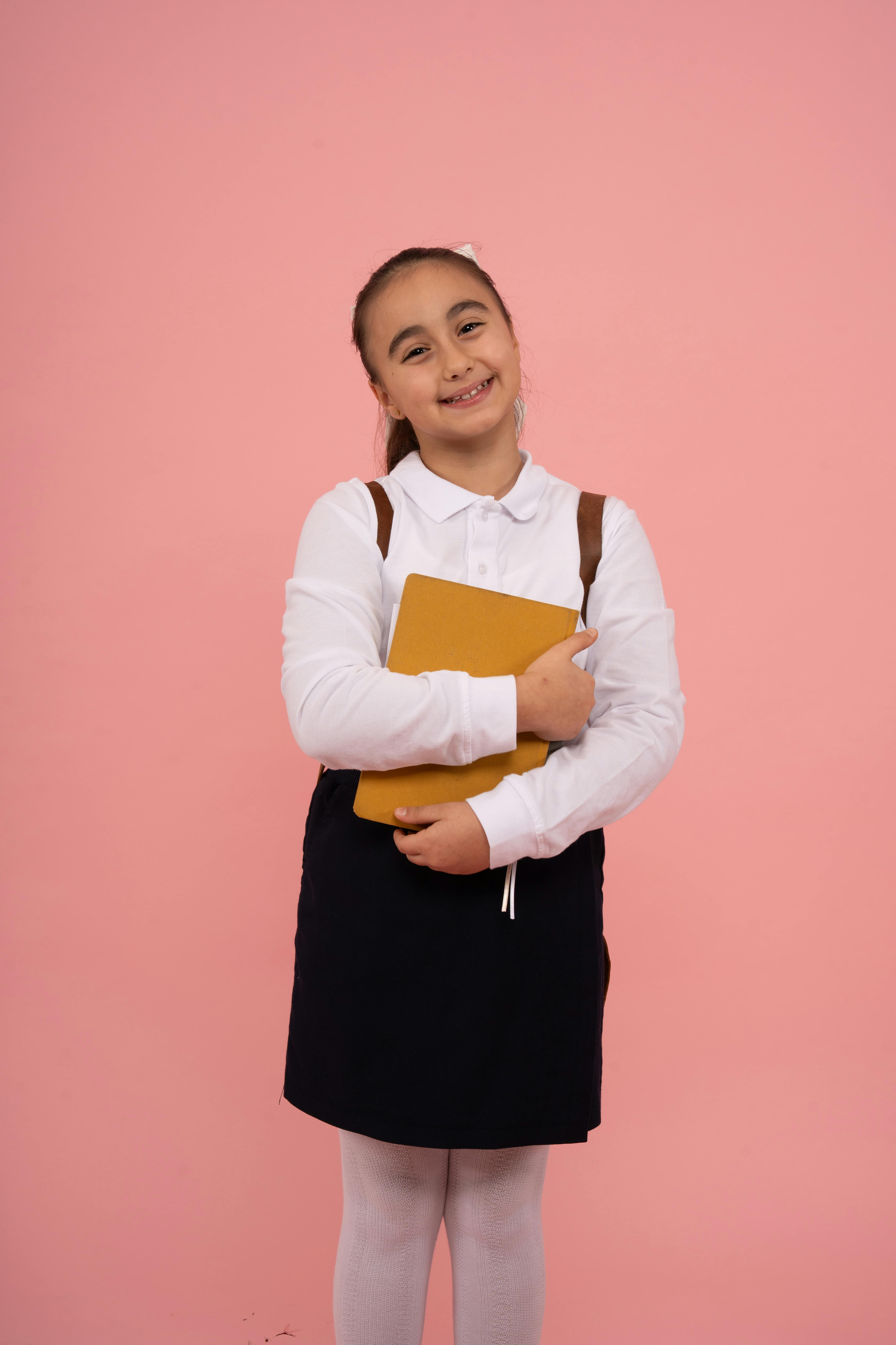 Cheerful schoolgirl in uniform holding a book, standing against a pink background.