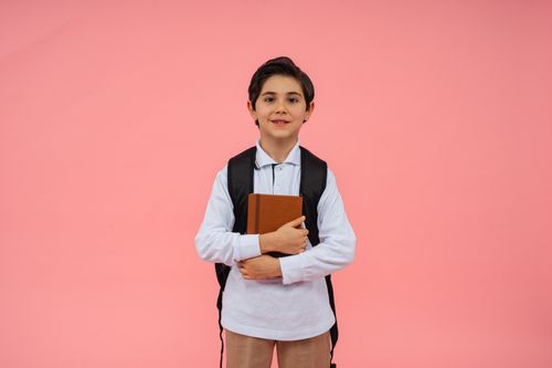 Free Young boy in school uniform holding a book with a pink background. Stock Photo