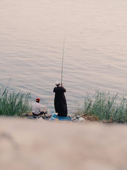 Two people fishing by the riverside during sunset, capturing tranquility.