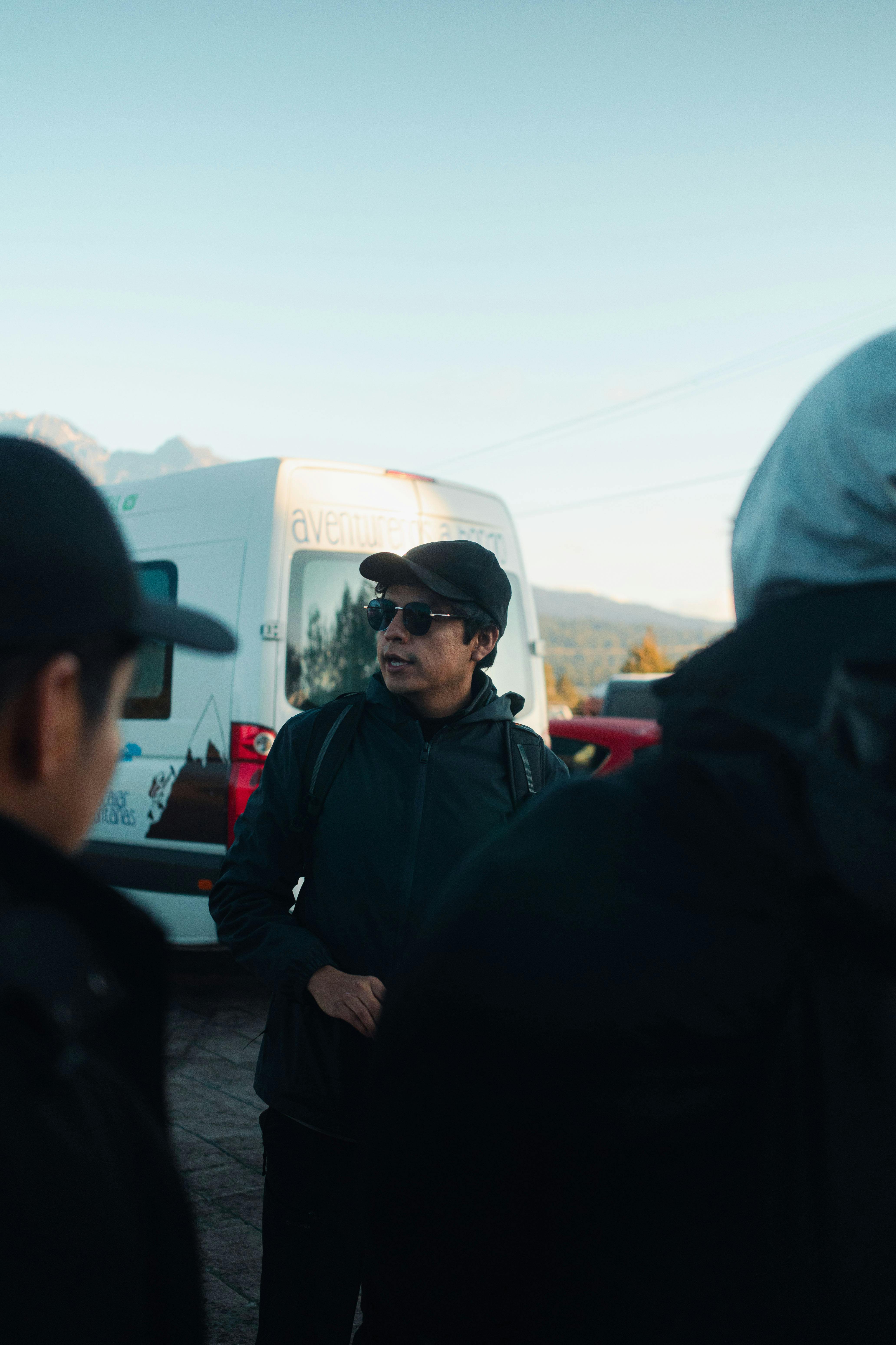 Three adults conversing by a parked van outdoors, dressed in casual attire.