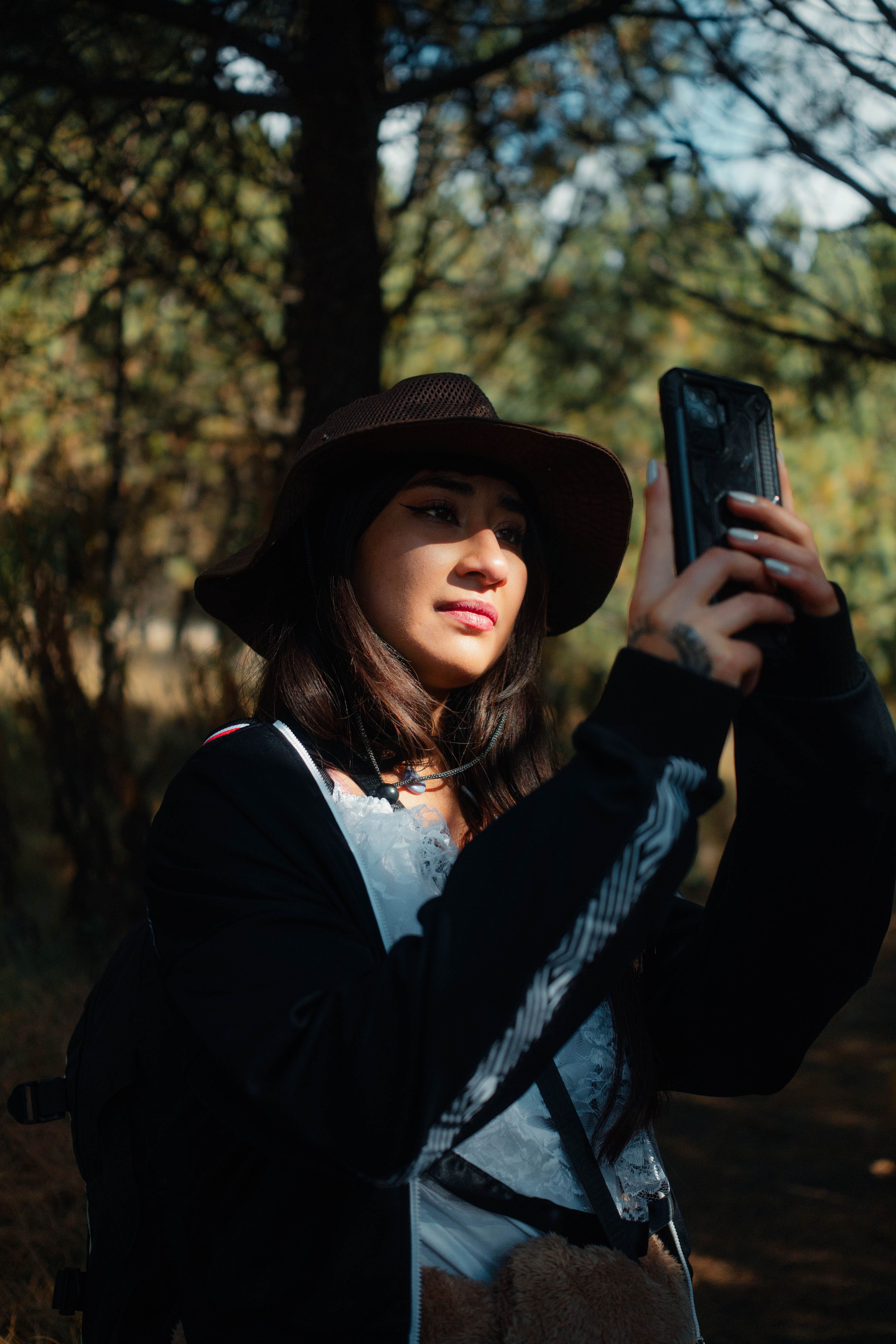 A young woman wearing a hat takes a selfie in a sunlit forest setting, capturing a serene outdoor mood.