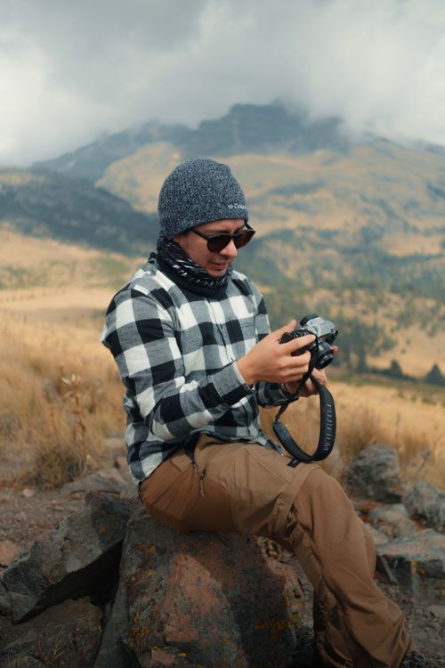 Free Man sitting on a rock capturing mountain scenery with a camera. Stock Photo