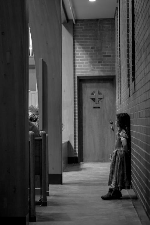 Grayscale Photo of Child in Dress Standing in Front of Door
