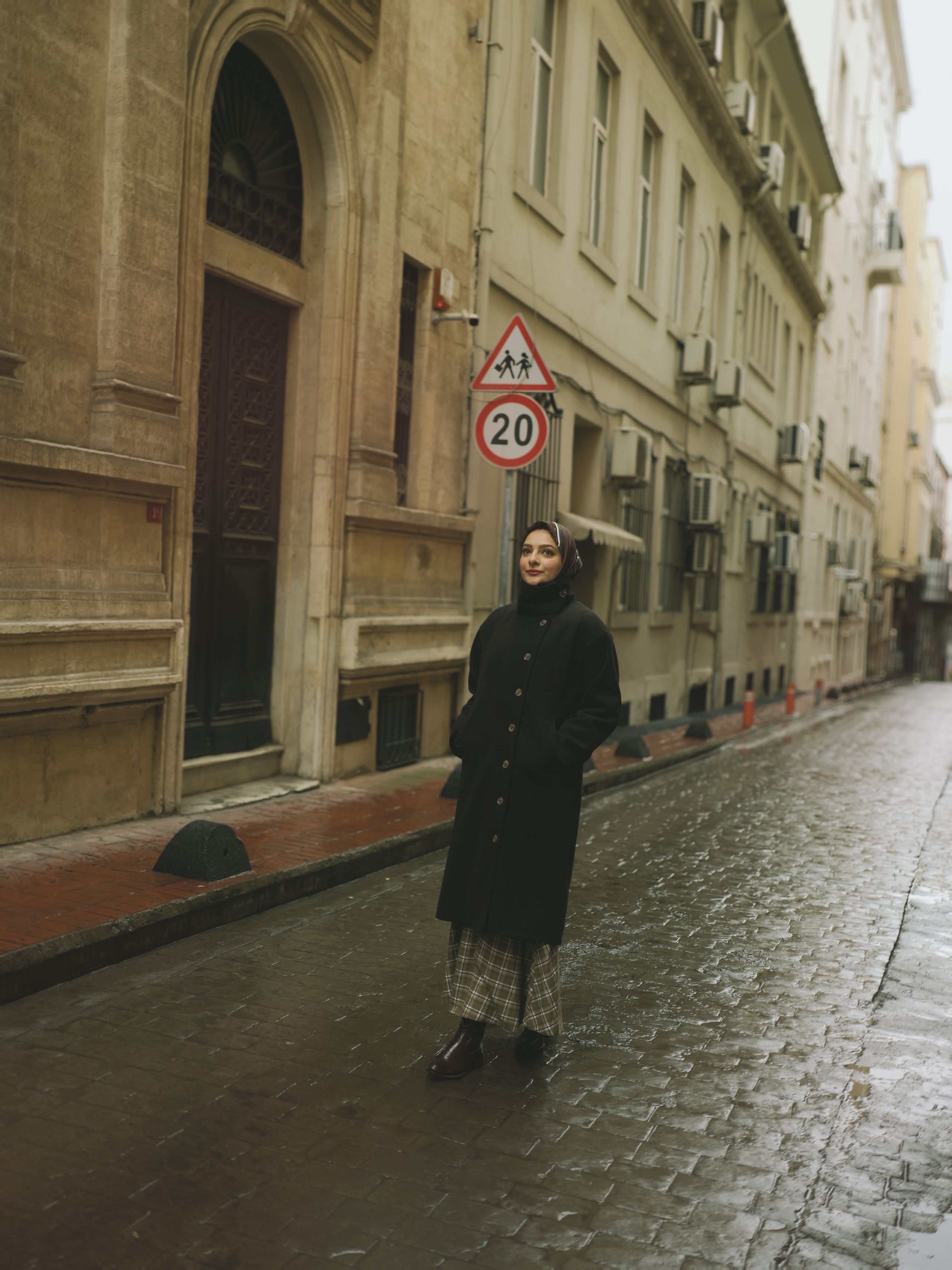 Woman in a long coat walking down a narrow, wet street.