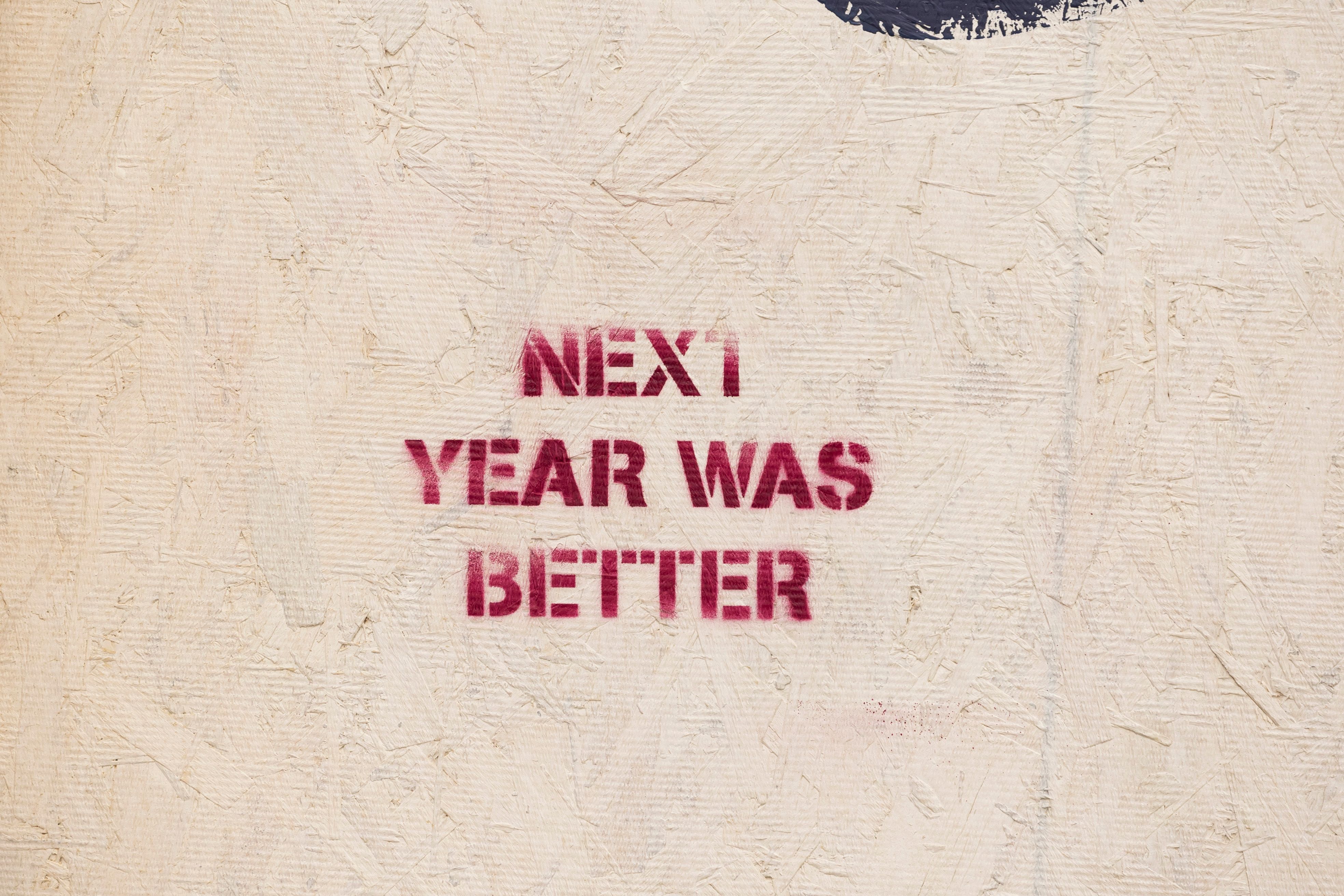 Rustic wall art with an inspirational message 'NEXT YEAR WAS BETTER' stenciled on a textured surface.
