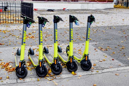 Five lime green e-scooters parked in a row on a leaf-strewn street during autumn.