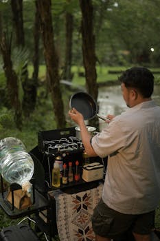 Man cooking on grill in lush West Java setting. Ideal for camping themes.