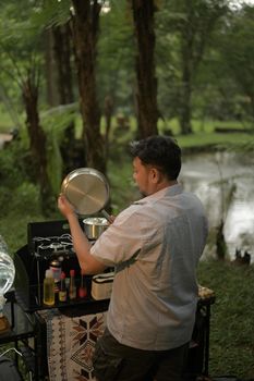 A man cooking outdoors by a scenic riverside in West Java, surrounded by lush greenery.