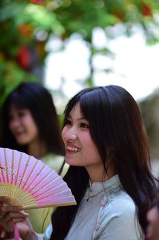 Smiling woman holds a colorful fan wearing traditional attire in a vibrant outdoor setting.