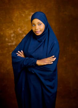 Portrait of a woman in a blue hijab against a warm brown backdrop, arms crossed confidently.