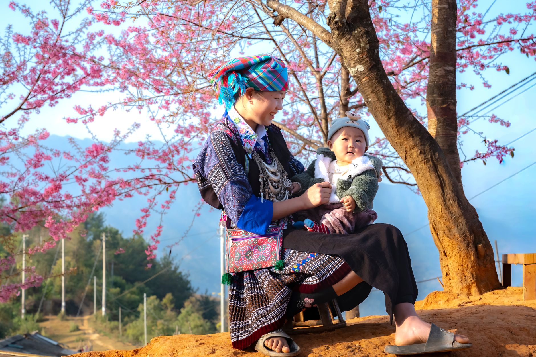 A mother and child in traditional attire sit under blooming cherry blossoms in a scenic outdoor setting.