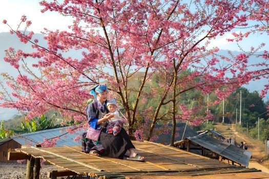 A mother and child sit under a vibrant cherry blossom tree in a rural setting, capturing a peaceful moment.