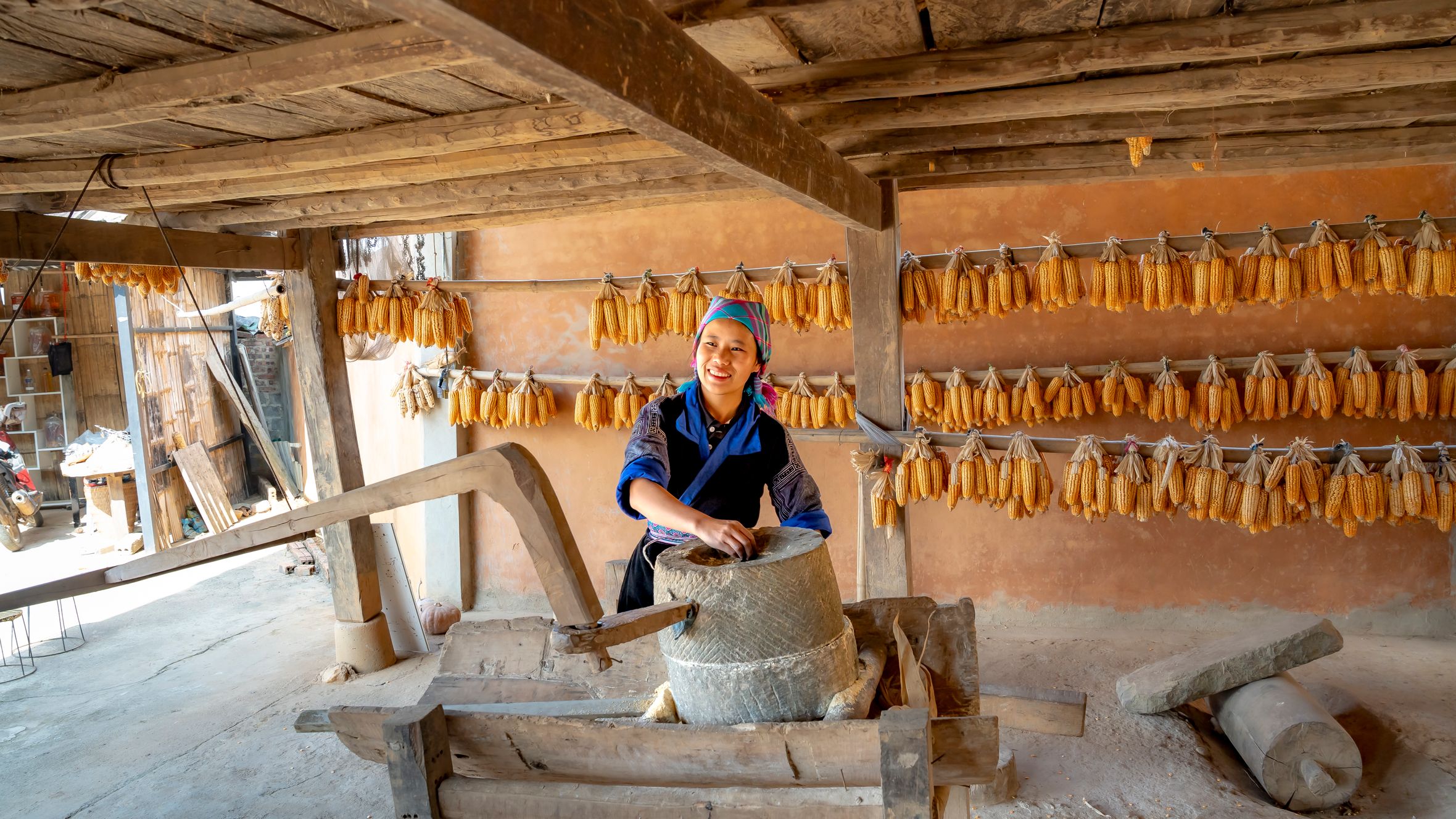 A woman using a traditional grinder in a rural setting with corn drying in the background.