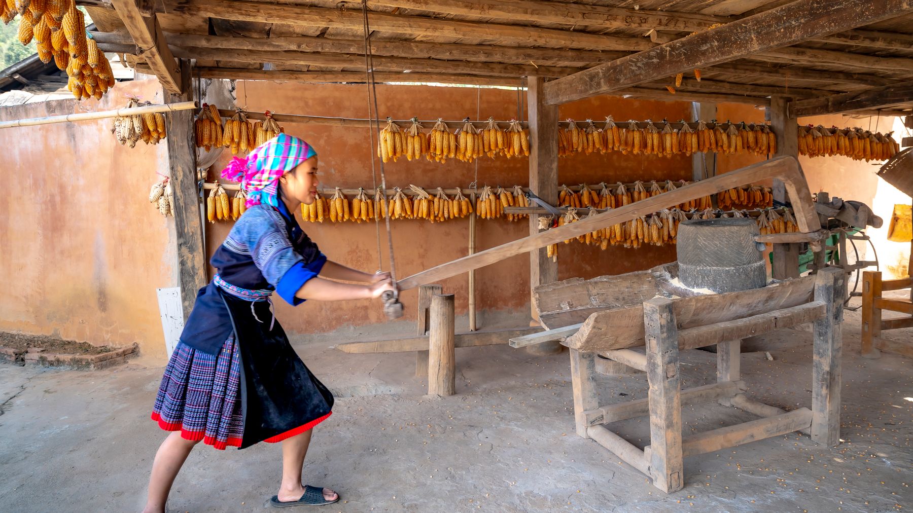 Woman using traditional corn milling tool in rural Vietnamese setting with hanging corn
