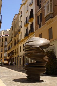 A unique sculpture stands in a sunny street surrounded by classic European architecture.
