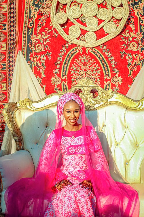 Free A woman in vibrant pink traditional dress sitting on a luxurious white and gold couch against a rich tapestry backdrop. Stock Photo