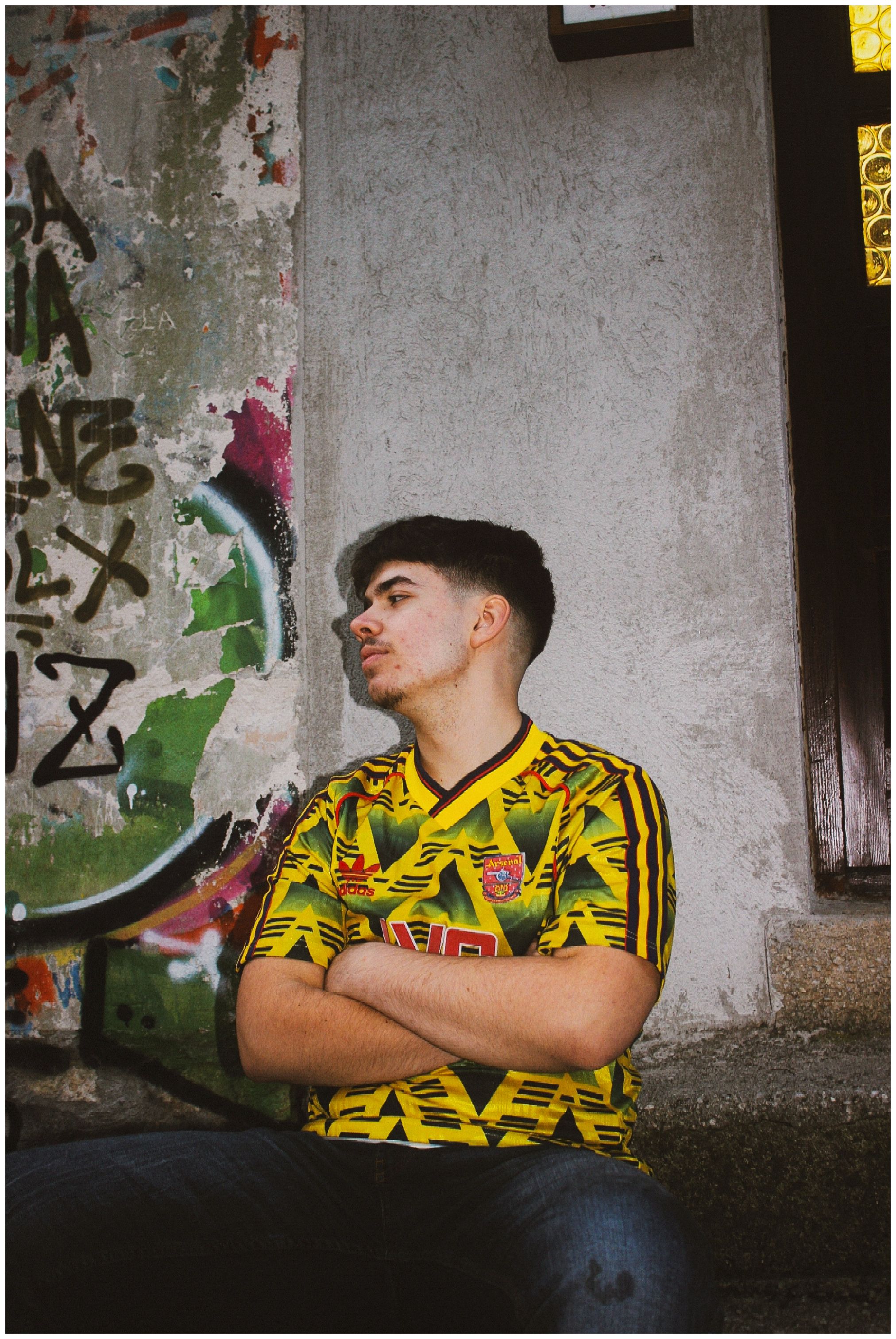 A young man in a colorful jersey poses confidently against a vibrant graffiti wall outdoors.