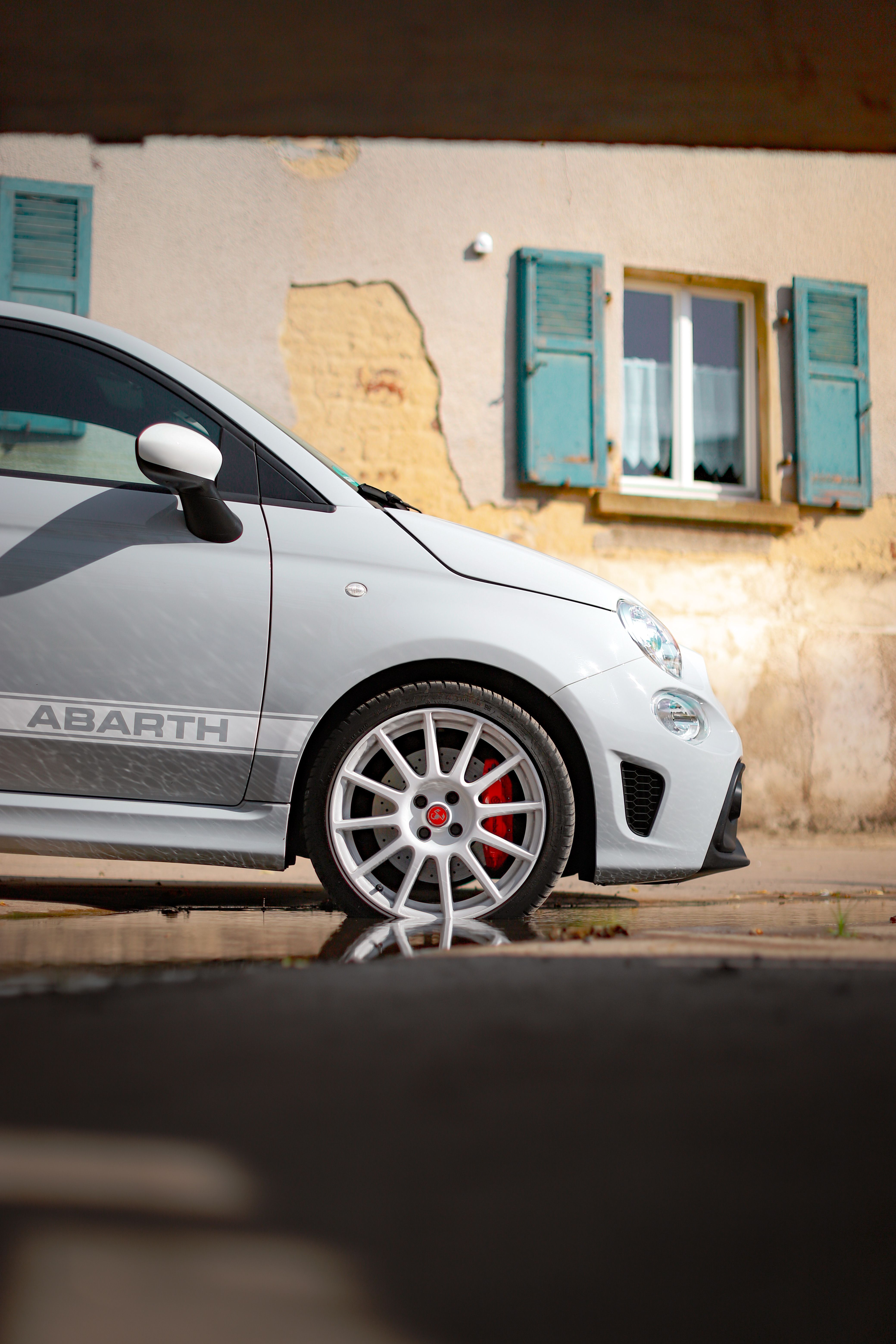 Bezpłatne Zbliżenie na samochód Abarth zaparkowany obok rustykalnego budynku z turkusowymi okiennicami. Zdjęcie z galerii