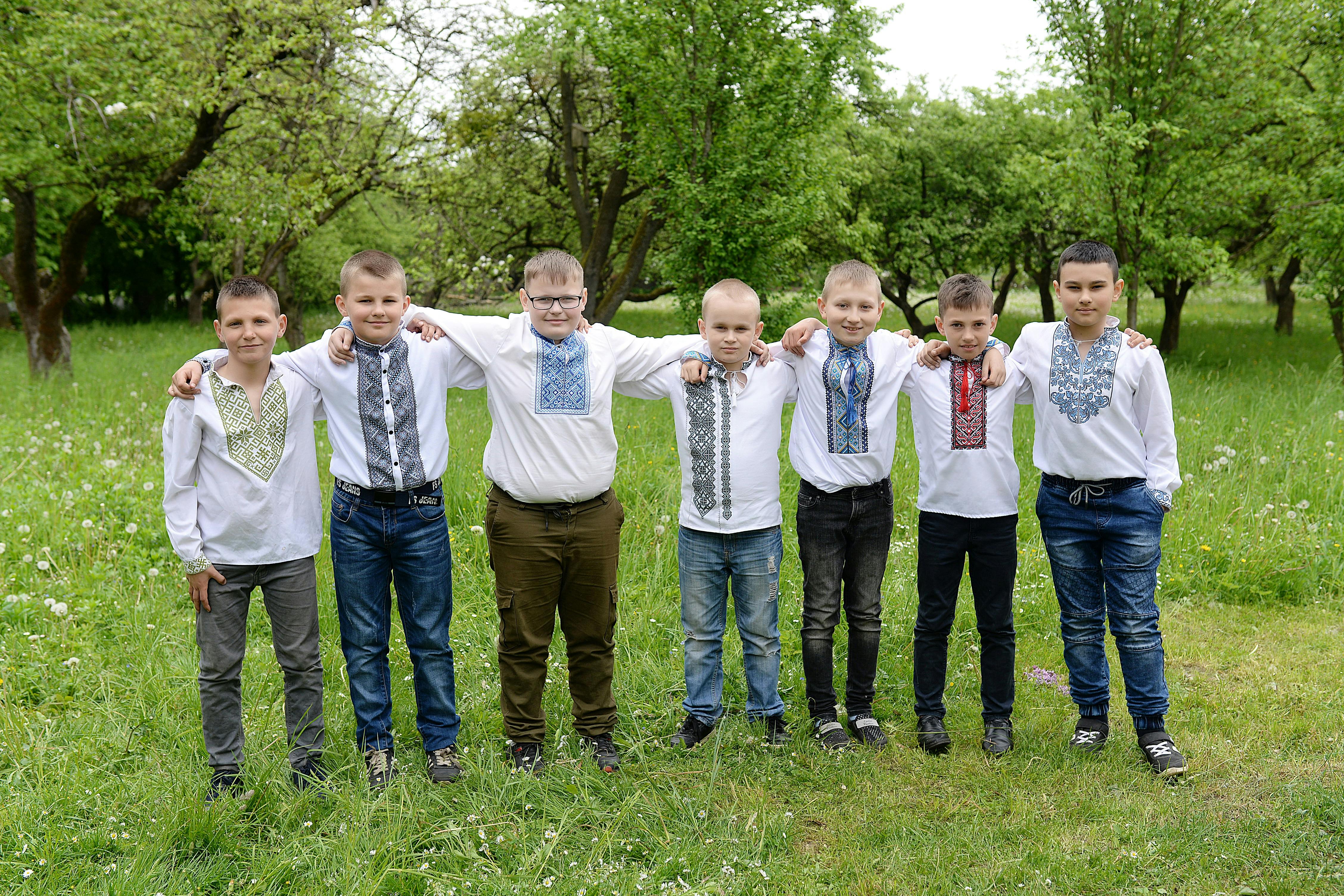 Seven boys wearing traditional embroidered shirts pose together in a green park.