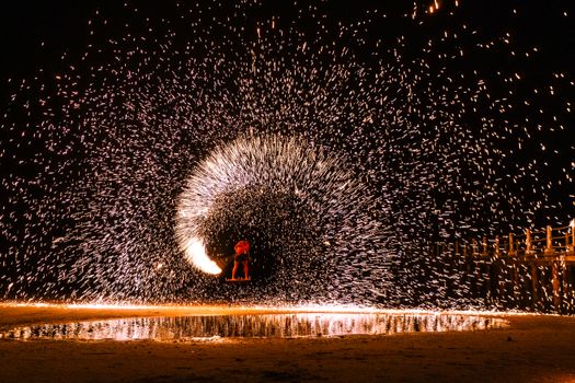 Dramatic long exposure shot of a fire performance with spinning steel wool creating a mesmerizing light effect on a beach at night.