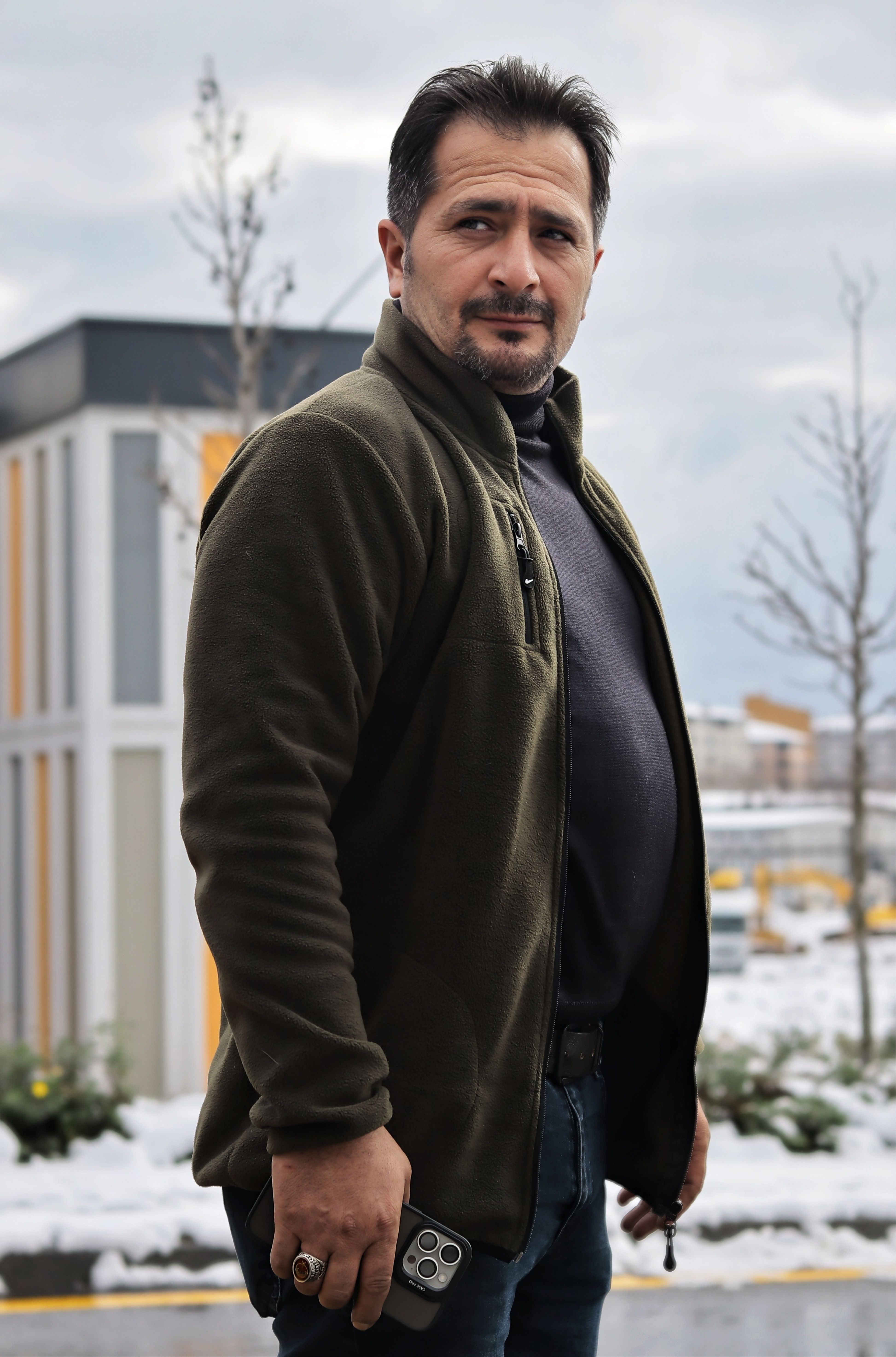 Man standing outdoors in winter, holding mobile phone, with snowy urban backdrop.