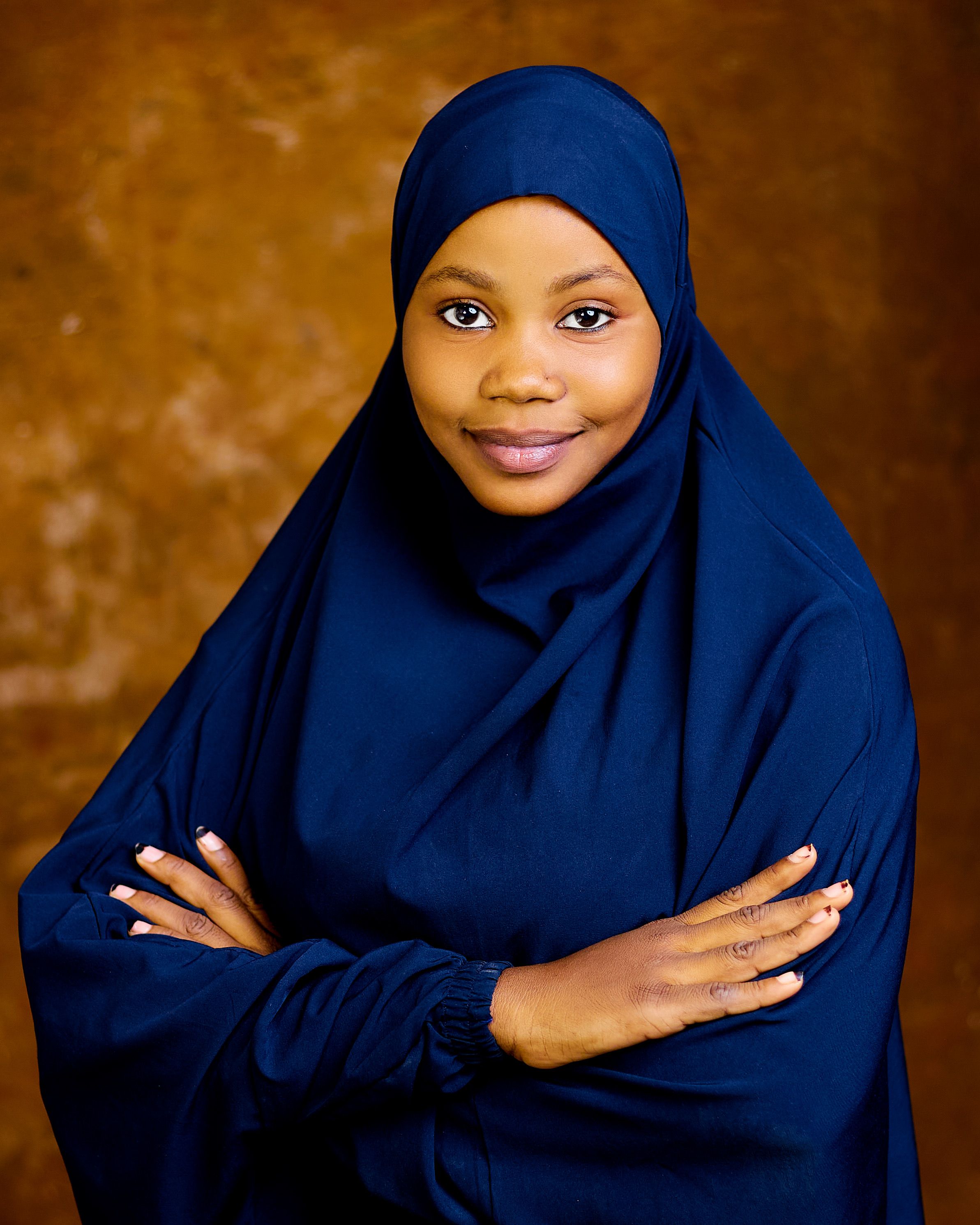 Portrait of a smiling woman in a navy blue hijab with crossed arms against a warm background.