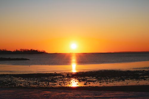 Free Serene sunset casting an orange glow over a calm ocean with a reflective shoreline. Stock Photo