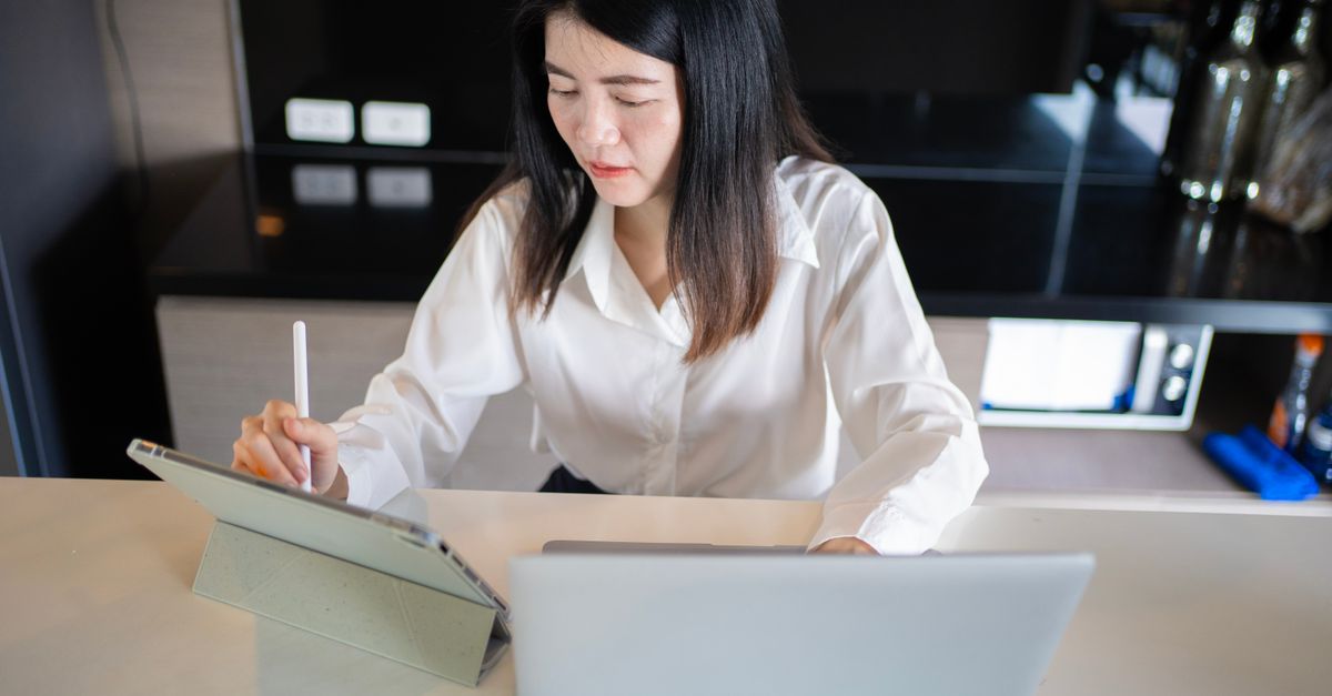 Asian woman multitasking with a laptop and tablet in a modern home office.
