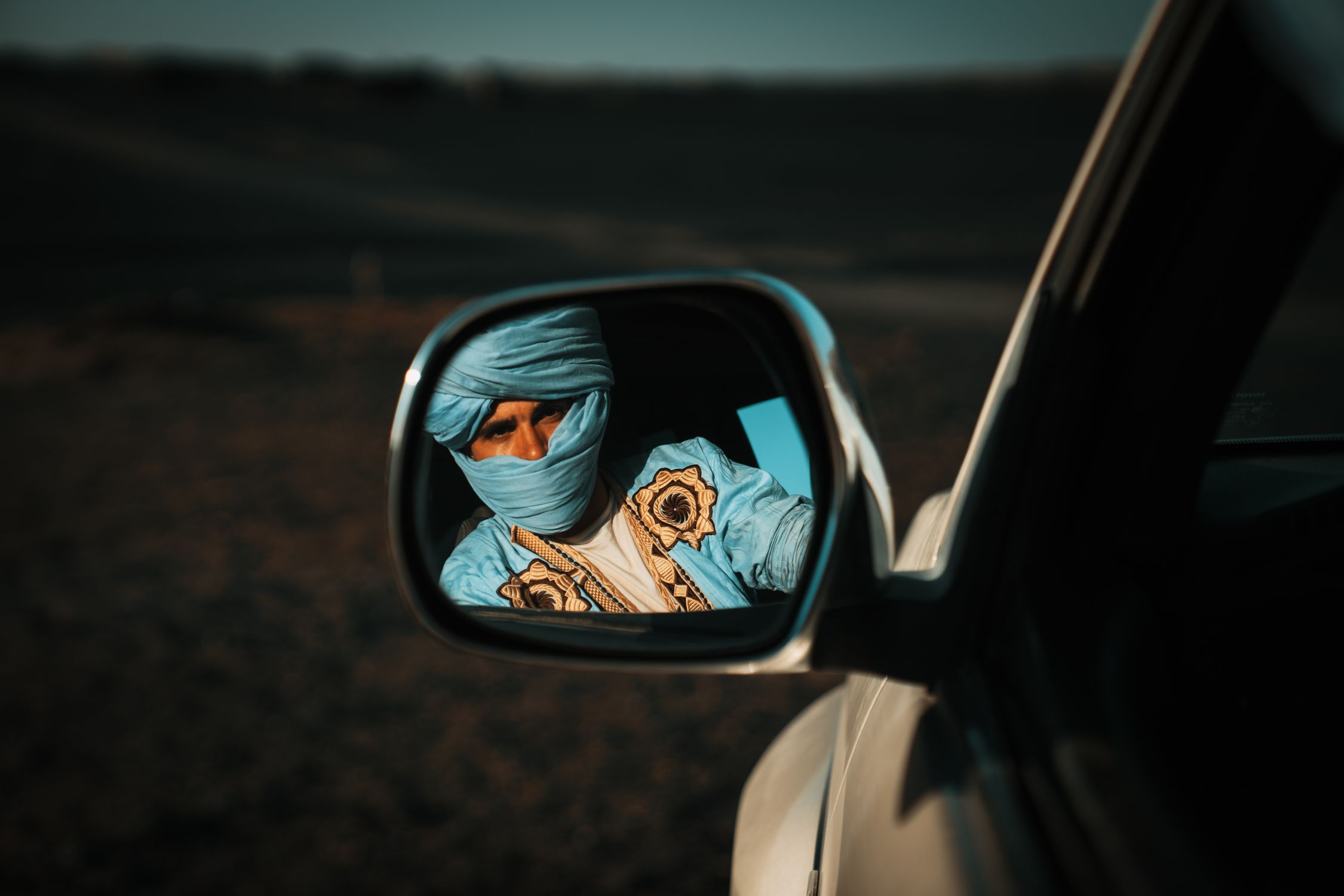 A man in traditional attire and turban is reflected in a car's side mirror, captured outdoors.